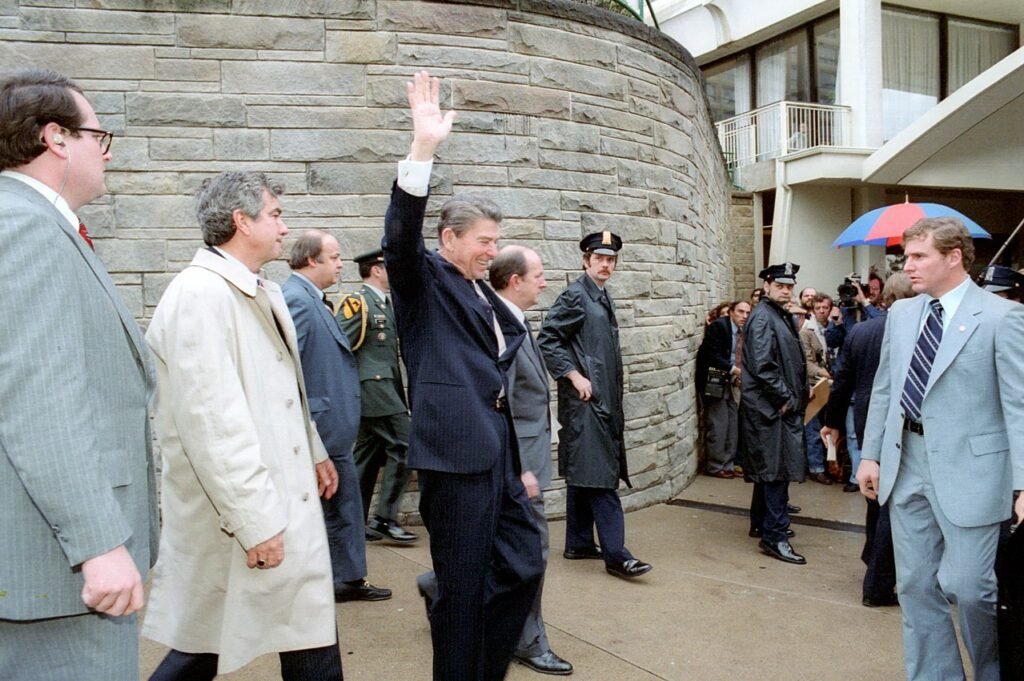 Ronald Reagan acena pouco antes de ser baleado. A partir da esquerda estão Jerry Parr , de sobretudo branco, que empurrou Reagan para dentro da limusine; o secretário de imprensa da Casa Branca , James Brady , gravemente ferido por um tiro na cabeça; assessor Michael Deaver ; um policial não identificado; o policial Thomas Delahanty , que levou um tiro no pescoço; e o agente do Serviço Secreto Tim McCarthy , que levou um tiro no peito.