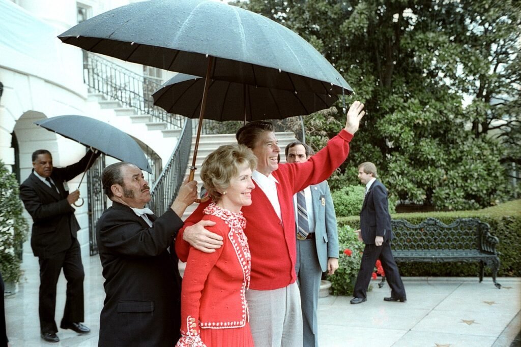 Reagan acena da Casa Branca após retornar do hospital em 11 de abril. Ele usava um colete à prova de balas sob o suéter vermelho.